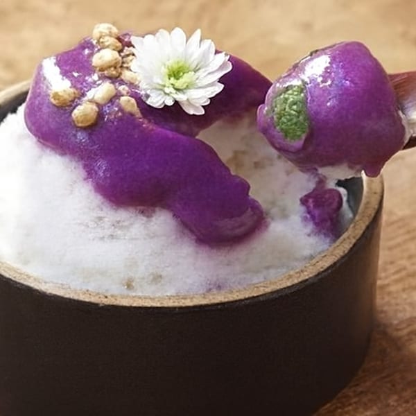 A dessert with purple topping, white flower, and nuts in a dark bowl on a wooden surface.