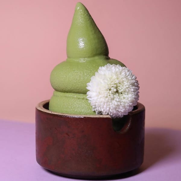 Green soft-serve ice cream in a brown ceramic cup, garnished with a white flower.