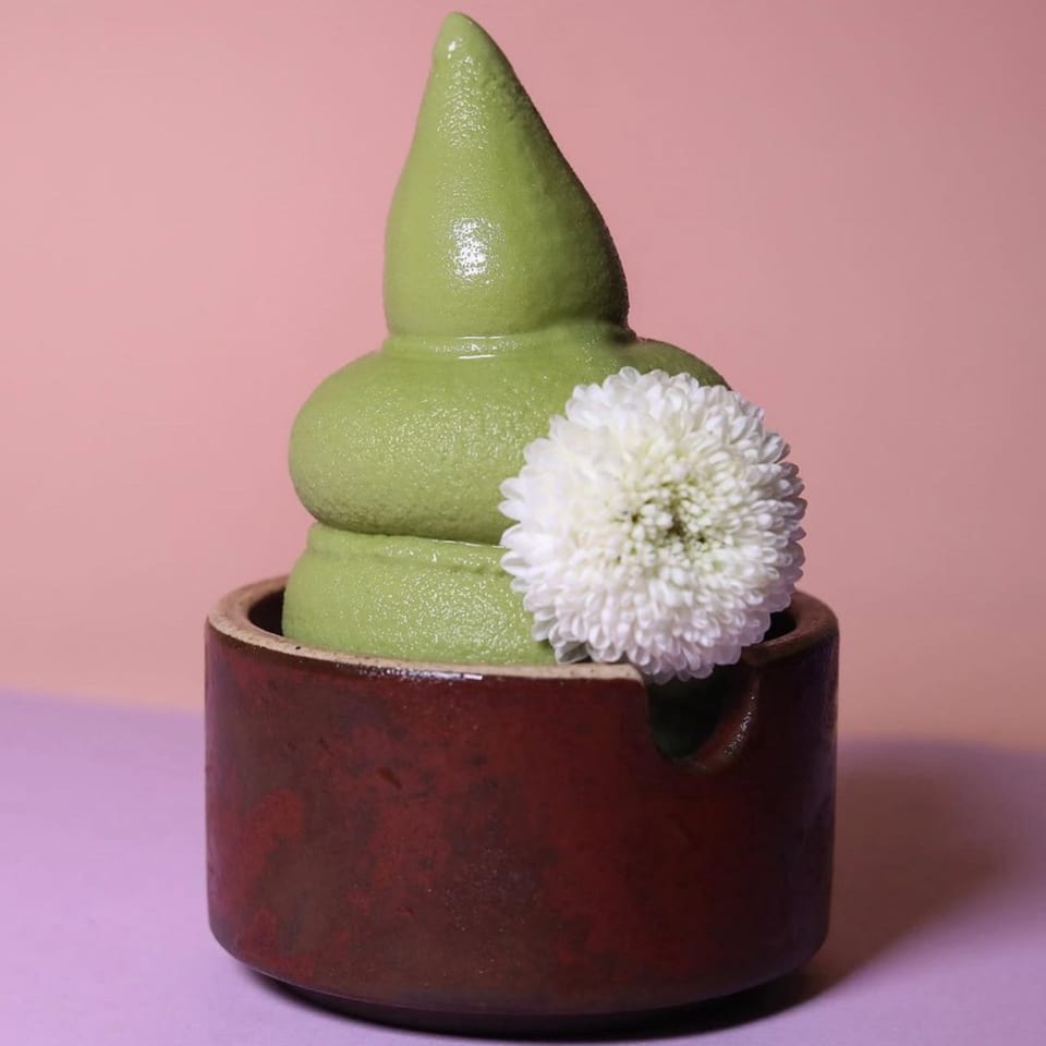 Green soft-serve ice cream in a brown ceramic cup, garnished with a white flower.