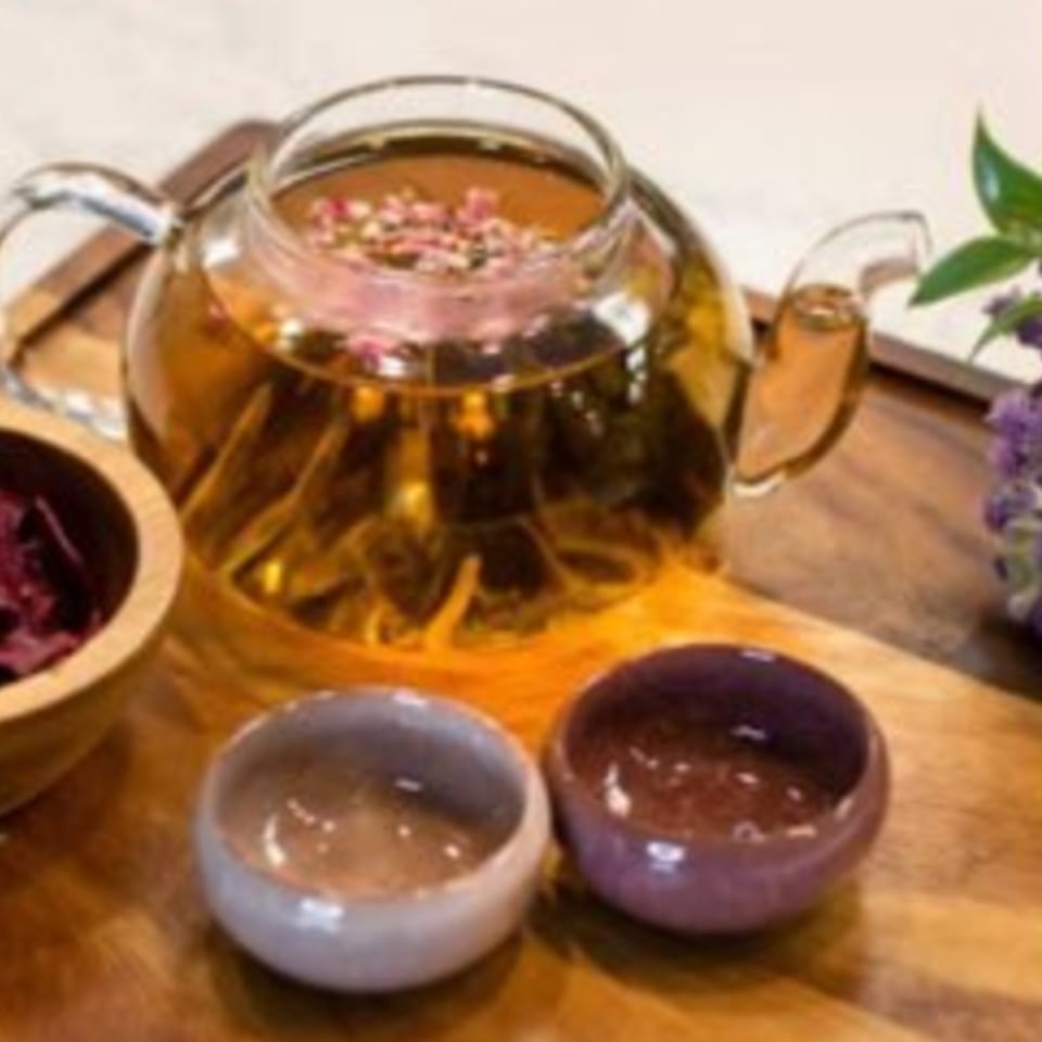 Glass teapot with rose petal tea, two small cups, and dried herbs on a wooden tray.