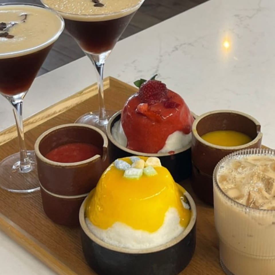 A wooden tray with two espresso martinis, two colorful shaved ice desserts, and two small sauce bowls.