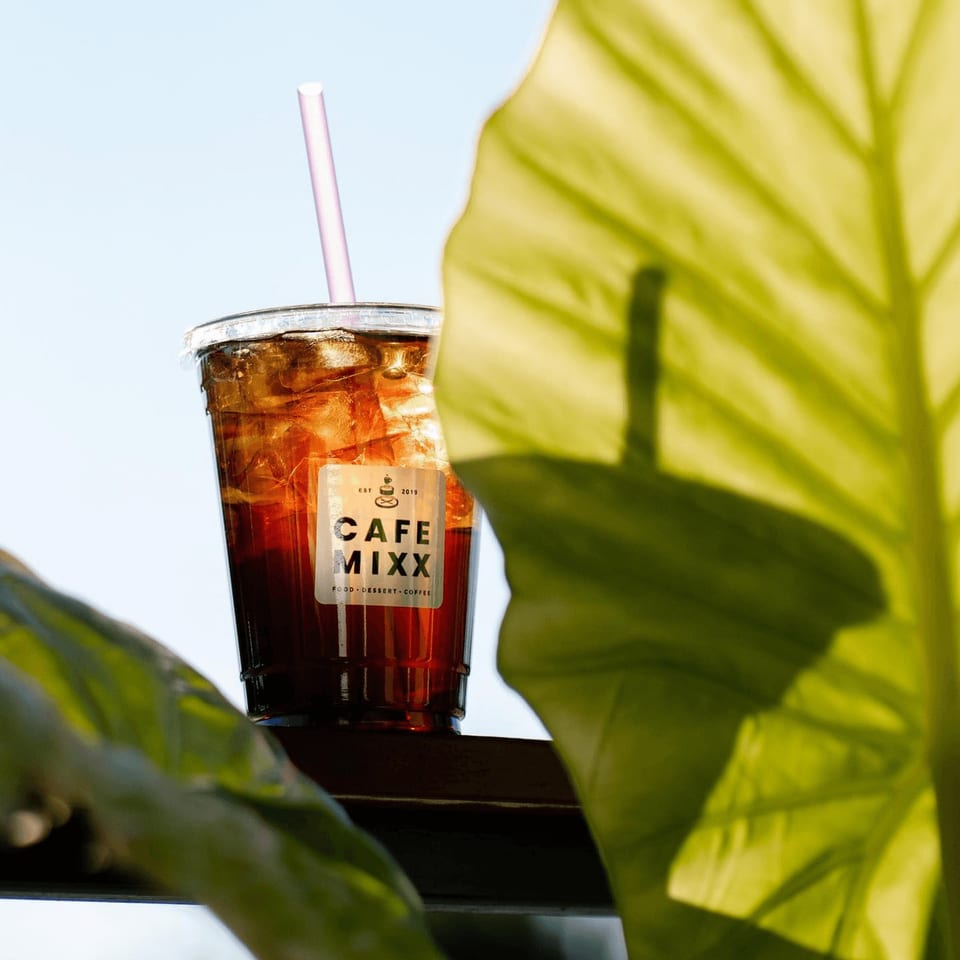 Iced coffee with a pink straw and Cafe Mixx logo, framed by green leaves against a bright sky.